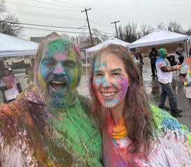 Two staff members smile with colorful paint on them.